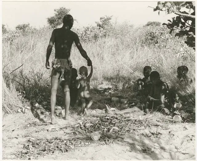 !Gai holding N!whakwe's hand, four people sitting, 1955. Peabody Museum